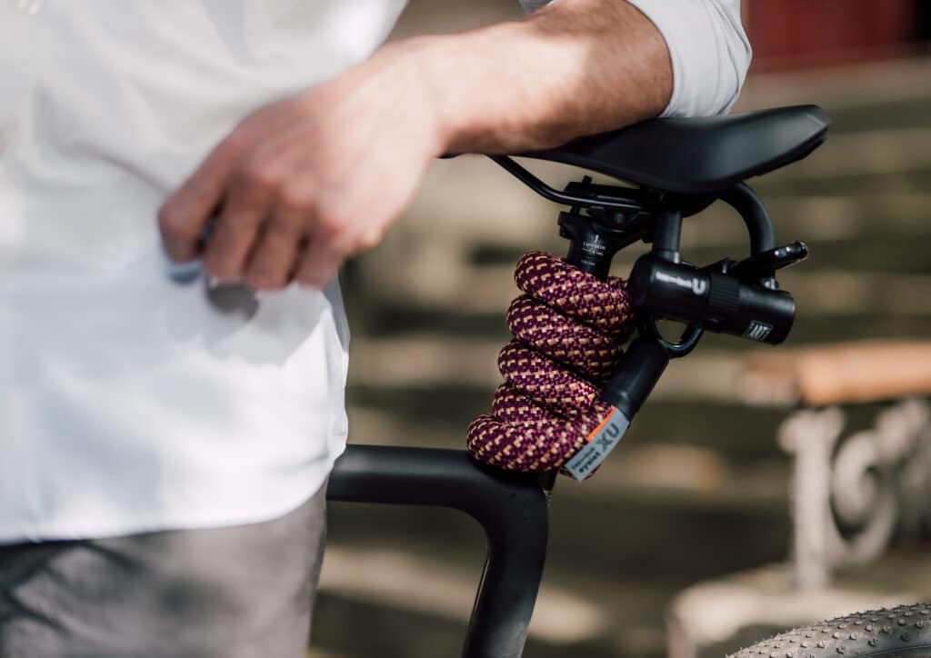 'tex–lock eyelet' in colour chateau red with U-lock are flexibly wrapped around a black bike's seat post, a male model leaning against it