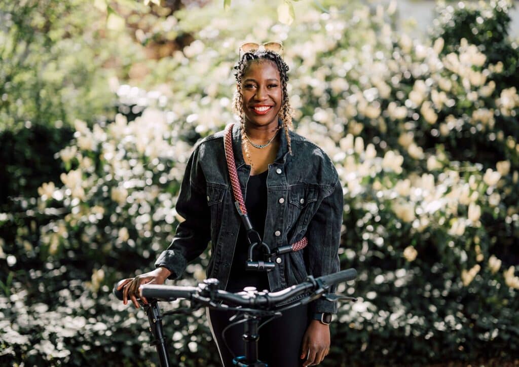 Female model smiling wearing 'tex–lock eyelet' in colour chateau red with U-lock around her shoulder and holding the bike saddle